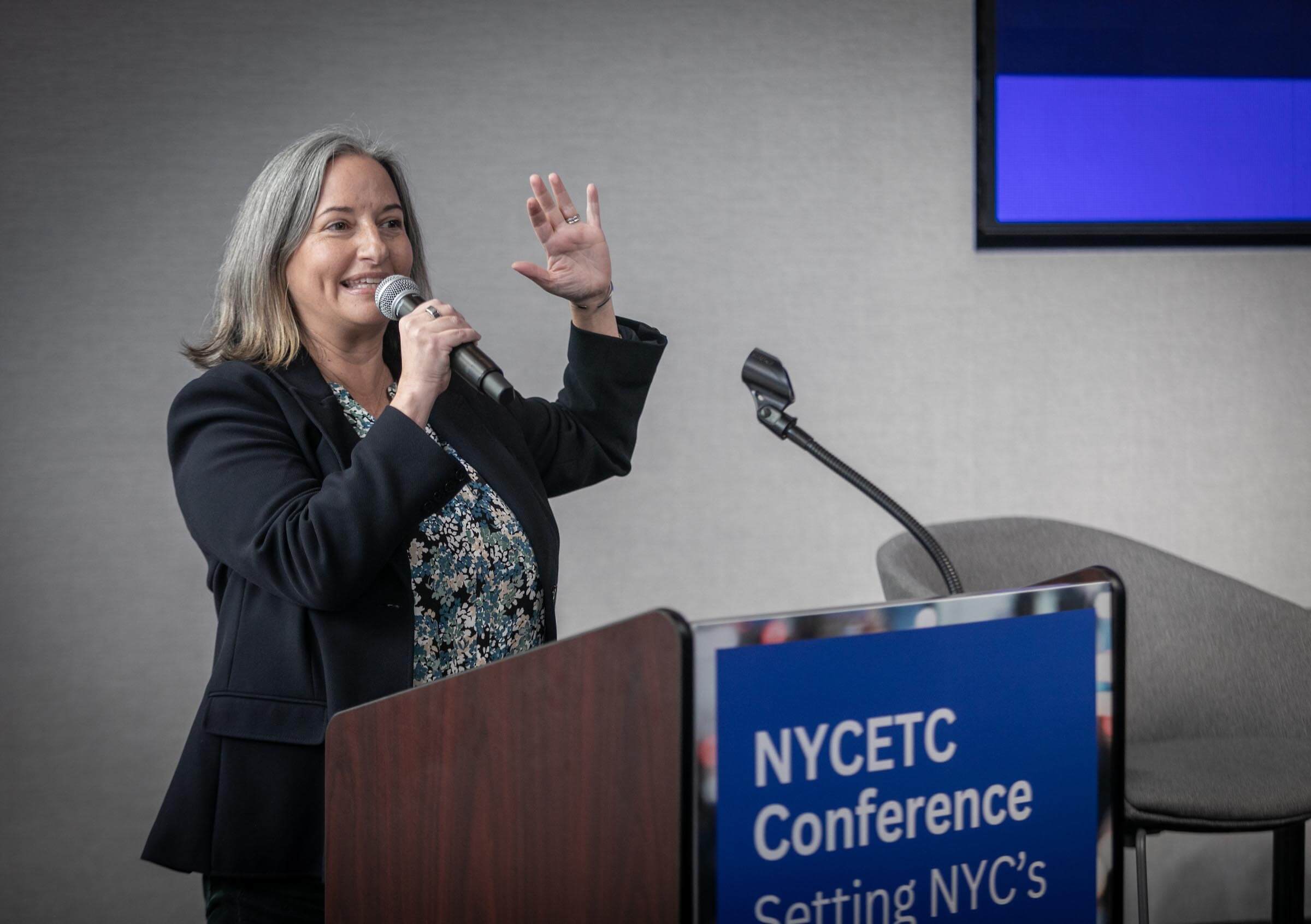 Jennifer Mitchell holds up a microphone to her mouth and her hand raised in the air at the podium at the 2024 NYCETC Conference