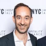 Andrew Rigie with very short light brown hair smiles in a slightly unbuttoned white dress shirt with a grey blazer.