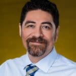 Anthony Barrows with with dark curly hair, goatee, and a light shirt with plaid tie, smiling warmly in front of a mustard yellow background.