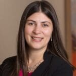 Ashley Putnam with long dark brown hair, and a small nose piercing, wearing a black blazer, magenta top, and pearl jewelry, smiling in front of a soft tan background.