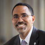 John B. King Jr. wearing a dark suit and light purple tie, smiling warmly in a softly lit setting.