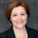 Christine Quinn with auburn hair in a black blazer, smiling in front of a dark textured background.