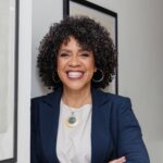 Dianne Morales smiling with curly hair in a blue blazer and white shirt wearing a gold necklace and hoop earrings.