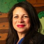 Eileen Torres with long dark hair and bright red lipstick, smiling in front of a colorful mural with trees