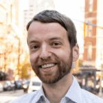 Eli Dvorkin with short brown hair and beard in a collared shirt, standing on a city street with blurred buildings in the background.