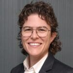 Erin Drinkwater with short curly hair, glasses, and a dark blazer, posed against a gray textured wall.
