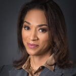 Jennifer Jones Austin with shoulder length brown hair smiling in a brown collared shirt and grey blazer.