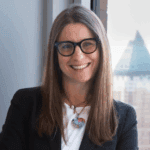 Julie Samuels smiling in front of a city view window, wearing glasses, a blazer, and a colorful heart-shaped necklace.