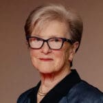 Kathryn Wylde wearing a navy blazer and dark glasses, smiling softly against a warm, tan background.