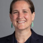 Kirstin Hill smiling warmly, wearing a navy blazer, green drop earrings, and a beaded necklace, with a neutral gray background.
