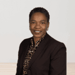Leah Rambo in a dark blazer and patterned blouse, smiling warmly in front of a light neutral background.