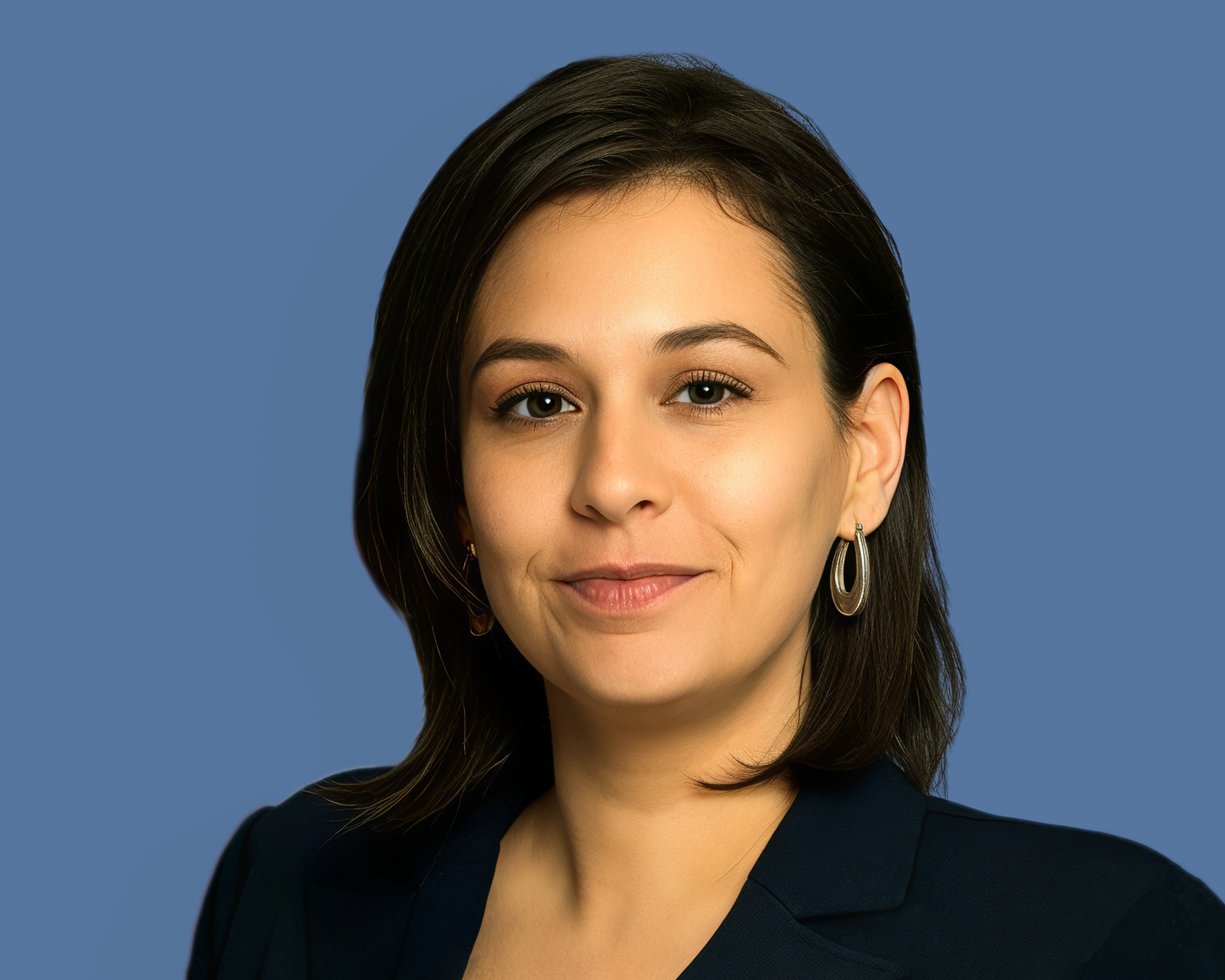 Liat Krawczyk, with shoulder-length dark hair, hoop earrings, and a navy blazer, posed in front of a blue background.