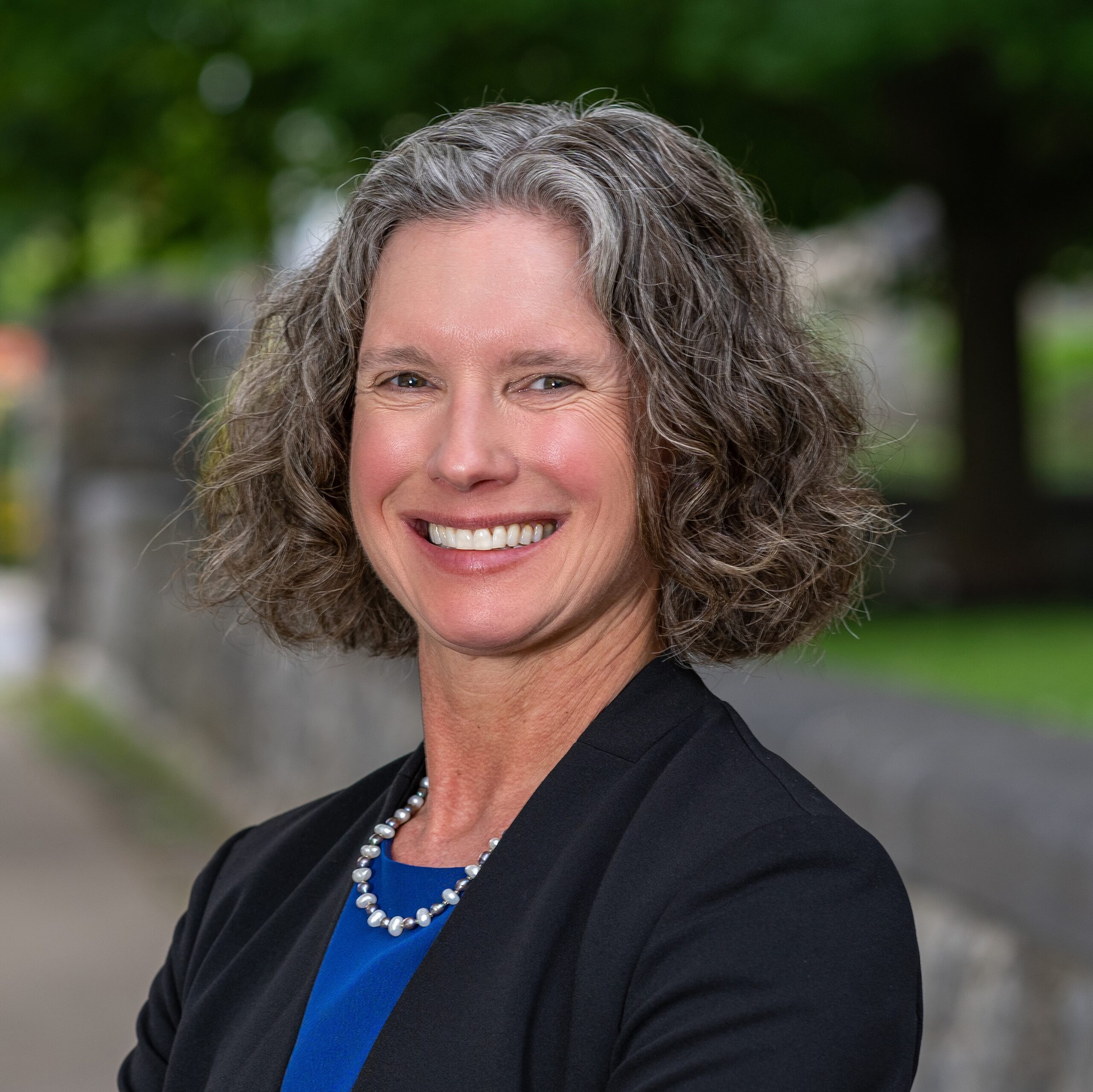 Lisa Vollendorf with wavy brown and grey hair in a black blazer and blue blouse, standing in front of greenery.