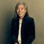 Maria Torres-Springer smiling with long black and grey hair in a dark velvet blazer and white top.