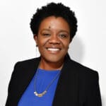 Michelle Rhone-Collins with short black hair smiling in a cobalt blue shirt and black blazer wearing a long gold necklace.