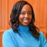 Pascale Leone with long black hair wearing a bright blue sweater, smiling in front of a wooden backdrop.