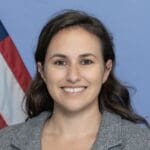 Rachel Lipson, smiling with long brown hair in a gray blazer, standing in front of a blue backdrop and part of an American flag.