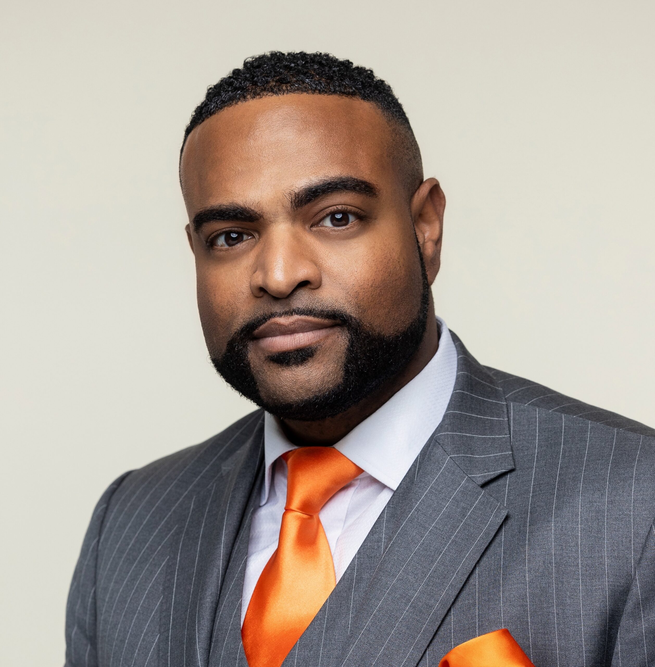 Randy Moore with short hair and trimmed beard in a pinstripe suit and bright orange tie, looking ahead with a neutral background.