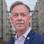 Randy Peers, wearing a navy blazer with a pin and standing in Industry City, Brooklyn.