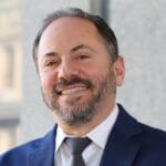 Robert Domanski, smiling with a salt-and-pepper beard and short hair, in a navy blue suit and gray tie, standing in front of a light stone building.