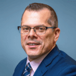 Scott French, smiling with a very short haircut, dark rimmed glasses, and a blue suit with a pink shirt and striped tie.