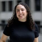 Shoshana Marder with long curly brown hair smiles in a black shirt with gold loop earrings.