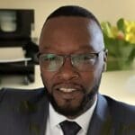 Terriell Peters, with a trimmed beard and close-cropped hair, smiles while wearing a dark suit, tie, and glasses.