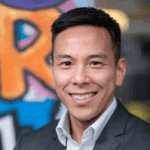 Kelsey Louie with short black hair wearing a grey suit in front a of graffiti covered wall.