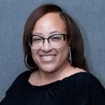 Alicia Johnson smiles in front of a grey backdrop, wearing black cat-eye glasses, large hoop earrings, and a black off-the-shoulder blouse.