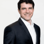 Darren Bloch with short dark hair and blue eyes, wearing a dark suit jacket over a white button-down shirt, smiling.