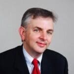 Doug Bauer, a man with short gray hair wearing a black blazer, light blue shirt, and red tie.