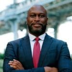 Gregg Bishop stands with arms crossed in a navy suit and red tie, smiling with arms crossed with a bridge in the background.