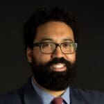 Marco A. Carrión slightly wearing glasses in a grey suit with a striped collared shirt and red tie