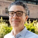 Jonathan Bowles smiles outdoors in front of a brick building, wearing glasses and a light blue button-down shirt.