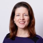 Katy Gaul-Stigge, with long brown hair, wearing a deep purple top with black accents, smiling.