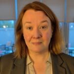 Merrill Pond wears a taupe blouse and dark blazer, standing indoors with warm lighting.