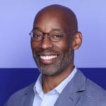 Michael Collins smiles in front of a two-tone purple background, wearing glasses and a light blue button-down shirt and jacket.