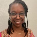 Rachel Cato smiles warmly in glasses and pink and gold top, wearing a gold “mama” necklace, nose ring, hoop earrings, and locs pulled back.
