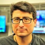 Ralph Ortega, with short dark hair and rectangular glasses, wearing a dark shirt and looking directly at the camera, with blurred TV screens in the background.