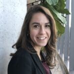 Rebekah Sides smiling, standing near a concrete wall and a large green plant, wearing a dark blazer and maroon top.