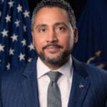 Robert Rodriguez with short curly hair and a trimmed beard, wearing a navy pinstripe suit, gray tie, and American flag lapel pin, stands in front of U.S. flag.