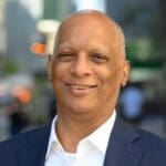 Roderick Jenkins smiles in a dark suit and open-collared white shirt, with a blurred urban background behind him.