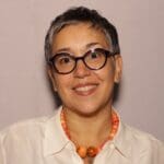 Sherazade Langlade wears round dark glasses, a white button-down shirt, and a bright orange beaded necklace, smiling against a plain mauve background.