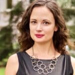 Stacy Woodruff smiles outdoors in front of a leafy background, wearing a black sleeveless top and a geometric statement necklace.