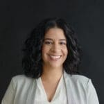 Tanzina Vega, with shoulder-length curly black hair, wearing a white blouse and light gray blazer, smiling.