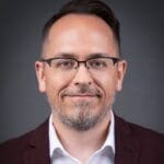 Gregory J Morris in dark rimmed glasses, short hair and a slightly gray short beard, and a burgundy blazer and white collared shirt.