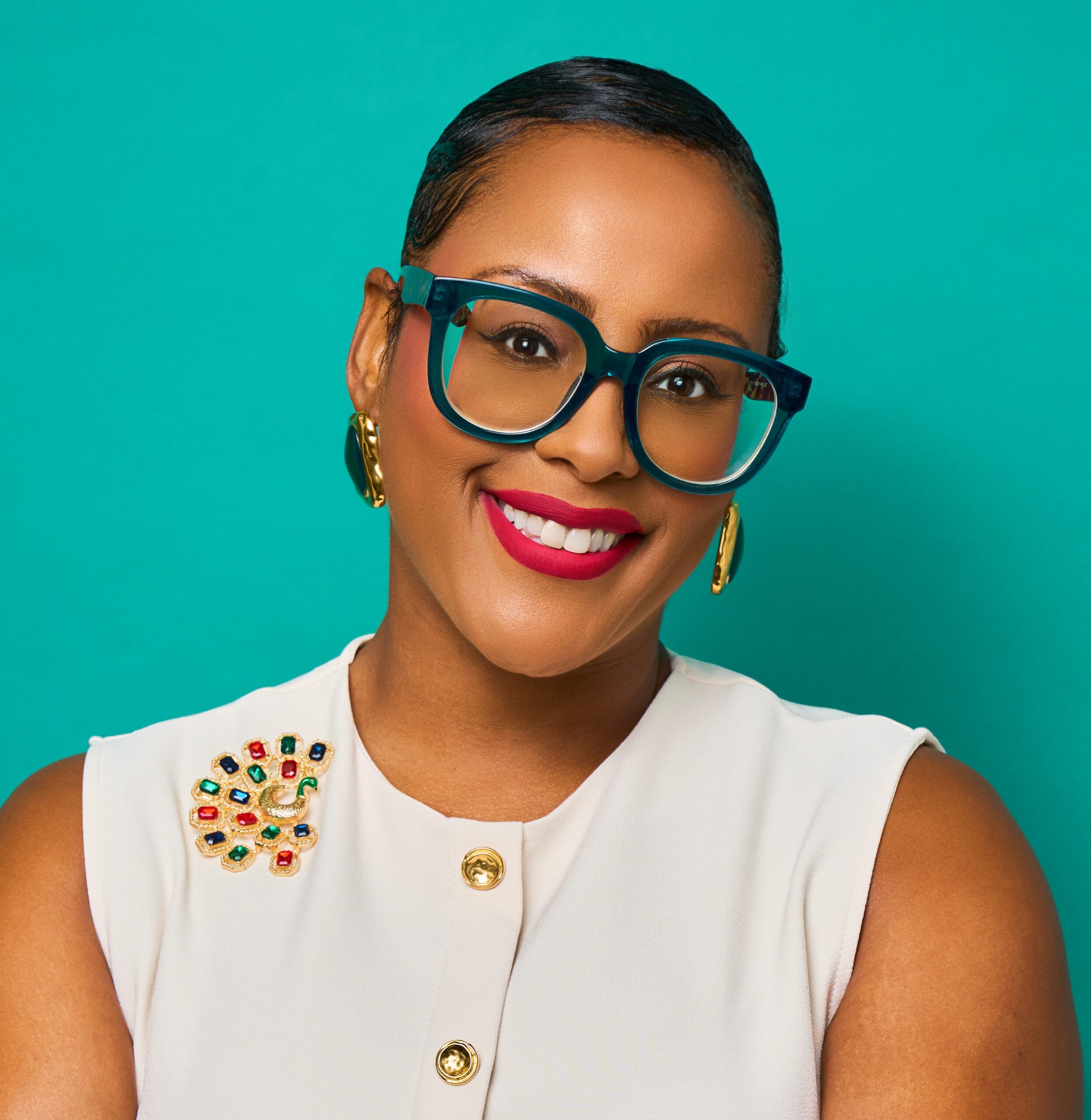 Kalilah Moon wearing teal thick glasses in a white short sleeved top with gold buttons and a multicolored golden pin on her lapel.
